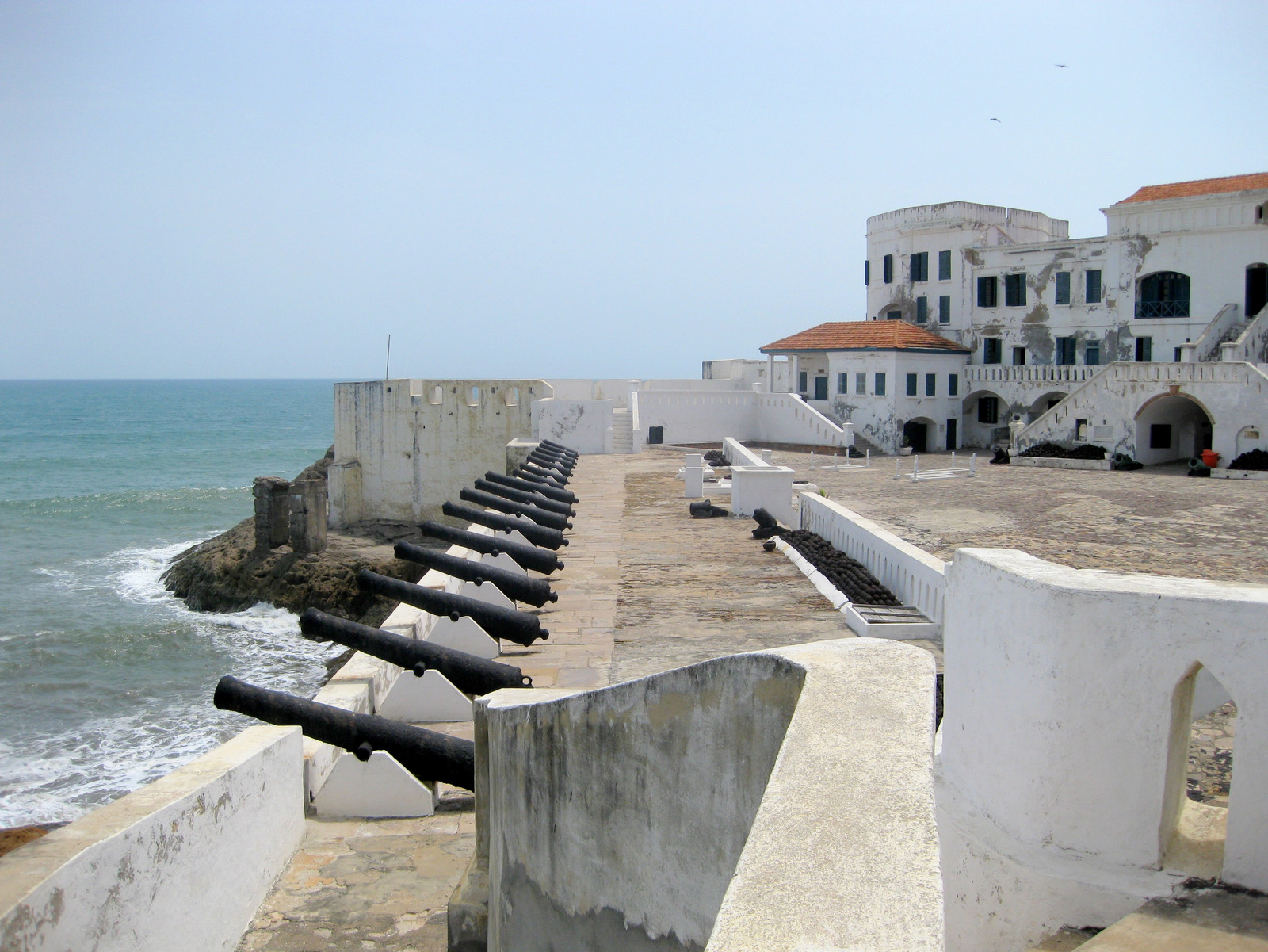 Cape Coast Castle's white coastal walls and courtyard, with a row of historic cannons facing the sea.